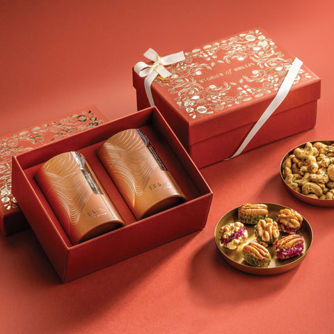 Red Stories of Bruijn gift box with white ribbon, containing two golden cylindrical containers and a bowl of Za’atar cashews alongside a bowl of stuffed walnut and pecan dried fruit wholes.