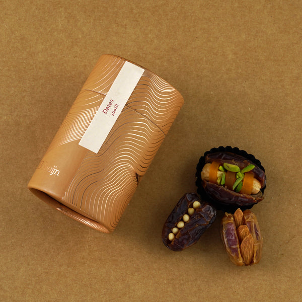 Golden cylindrical container of Bruijn Assorted Stuffed Dates placed beside dates filled with cashew nuts, almonds, mango peel, macadamia nuts, and candied orange peel.