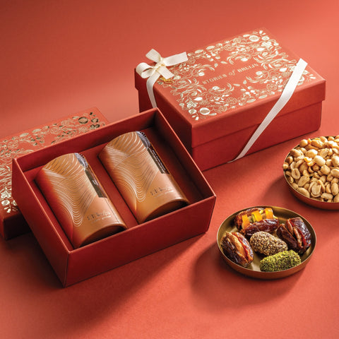 Red Stories of Bruijn gift box with white ribbon, containing two golden cylindrical containers and bowls of black truffle peanuts and assorted stuffed Mejdool dates.