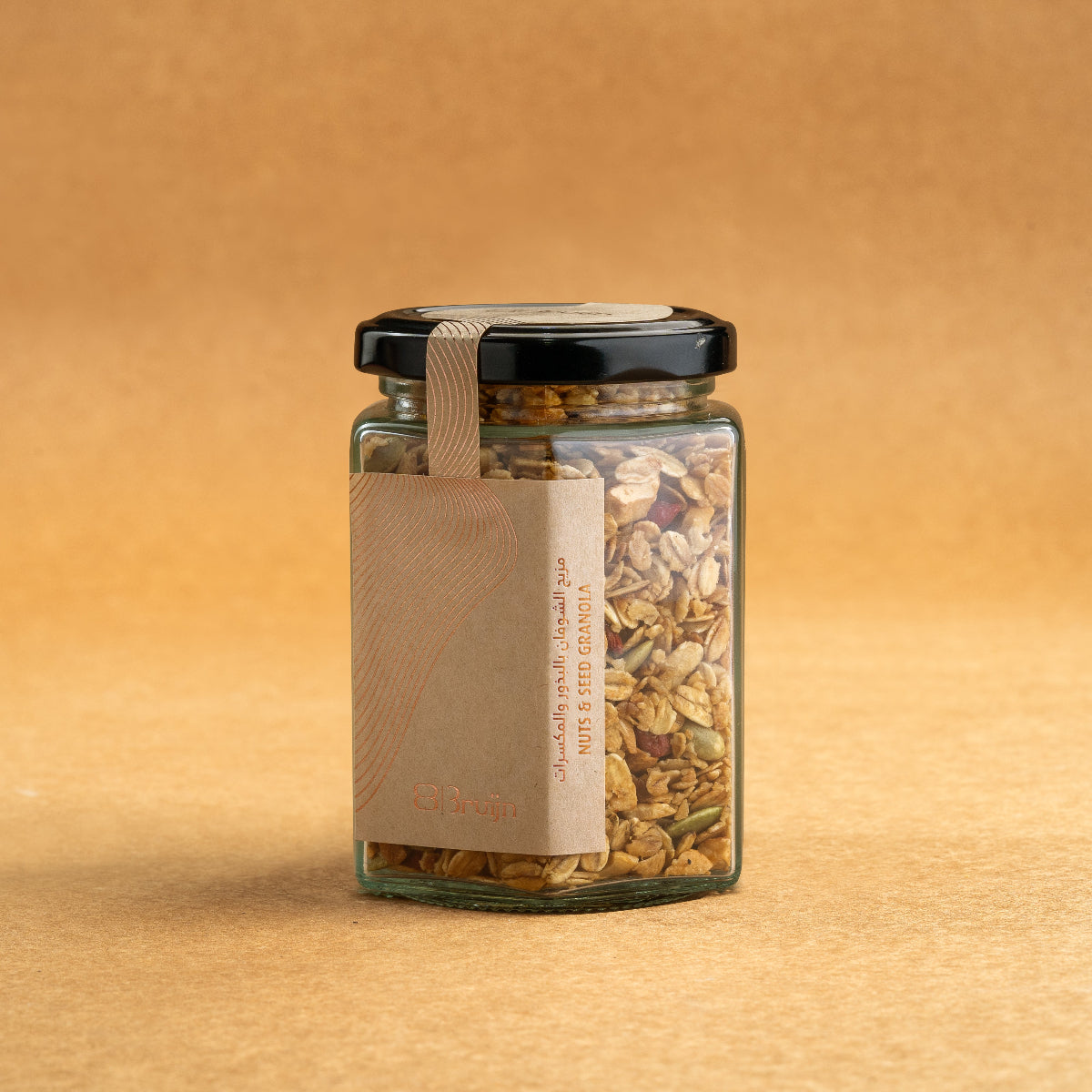 Glass jar of Bruijn Granola with black lid and kraft label, filled with rolled oats, almonds, hazelnuts, pumpkin seeds, and goji berries mixed with honey.