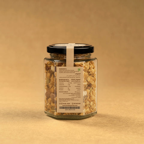 Glass jar of Bruijn Granola with black lid and kraft label, filled with rolled oats, almonds, hazelnuts, pumpkin seeds, and goji berries mixed with honey.