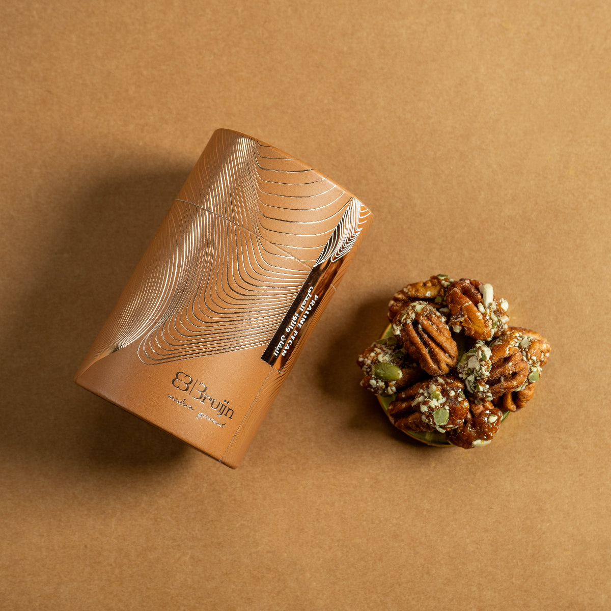 Brown cylindrical container of Bruijn Praline Pecans with minimalist design, holding caramel-coated pecans encrusted with roasted seeds for a sweet, crunchy bite.