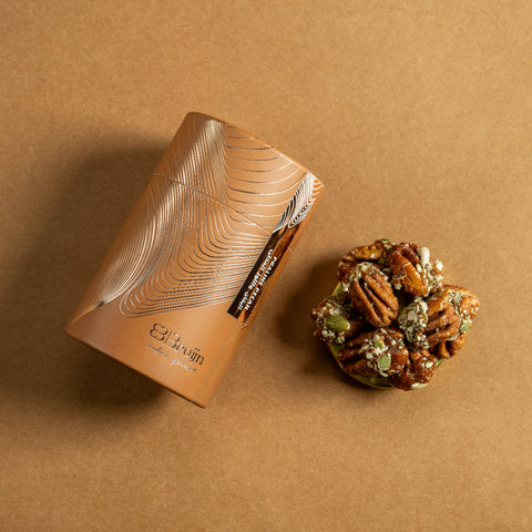 Brown cylindrical container of Bruijn Praline Pecans with minimalist design, holding caramel-coated pecans encrusted with roasted seeds for a sweet, crunchy bite.