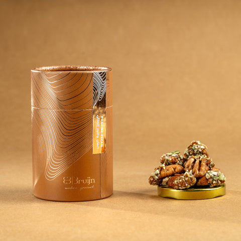 Brown cylindrical container of Bruijn Praline Pecans with minimalist design, holding caramel-coated pecans encrusted with roasted seeds for a sweet, crunchy bite.