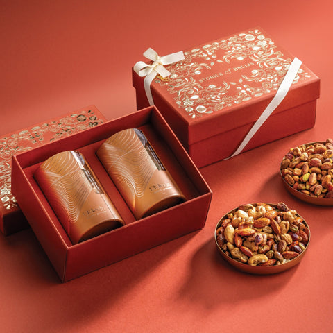 Red Stories of Bruijn gift box with white ribbon, containing two golden cylindrical nut containers, accompanied by bowls of salted caramel nut mix and lime & chilli pistachios.