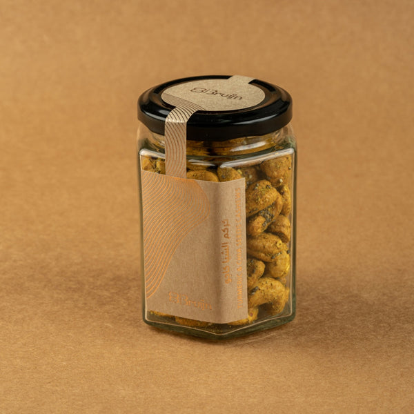 Glass jar of Bruijn Turmeric & Chia Seed Cashews with black lid and kraft label, filled with golden turmeric-coated cashews speckled with chia seeds.