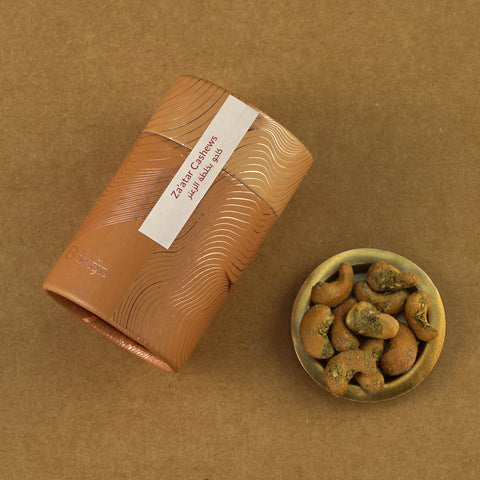 Golden cylindrical container of Bruijn Za’atar Cashews placed beside a small bowl of spiced cashews.