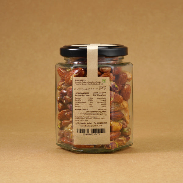 Glass jar of Bruijn Salted Caramel Nut Délice with black lid and elegant kraft label, filled with cashews, almonds, and pistachios coated in golden caramel.